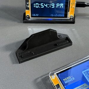 以下が含まれることがあります： 小型電子機器用の黒い3Dプリントスタンド。画面には時刻と日付が表示されます。QRコード付きの別のデバイスも表示されます。スタンドは長方形のベースと湾曲した上部を備えています。