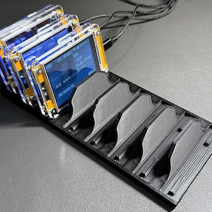 May include: A stack of small electronic devices with clear plastic cases and blue screens displaying data. They are mounted on a black holder with multiple slots. Wires extend from the devices.