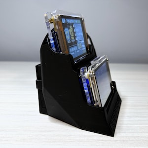 May include: A black, angled stand holding two electronic devices with screens. The screens display blue and white graphics. The stand is made of a matte black material and sits on a light-colored surface.
