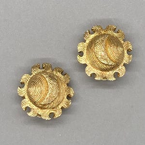 May include: A pair of circular gold-toned earrings featuring a textured, domed center with a raised, swirling sunburst pattern around the perimeter. The metal surface has a brushed finish, creating a distinct contrast between the central dome and the outer decorative edge of each earring.