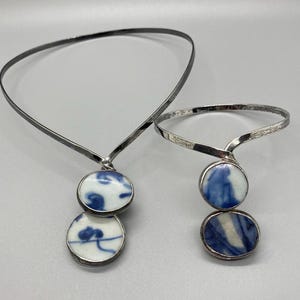 May include: A silver-toned metal choker necklace and a matching cuff bracelet, each featuring two circular ceramic pendants. The pendants are white with blue painted patterns, set in silver-toned metal frames that connect to the bands.