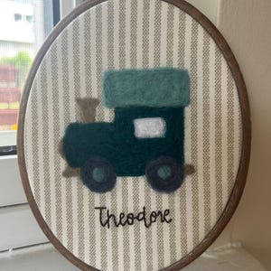 May include: An oval-shaped, framed embroidery featuring a green train design. The train is made of felt and has a white window. The background is striped, and the name "Theodore" is embroidered below the train. The frame is made of wood.