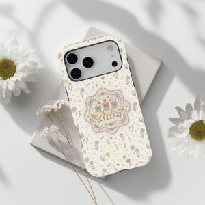 May include: A cream-coloured phone case with a floral pattern and a central design featuring three geese with blue and pink bows. The case has a black border and a camera cutout.