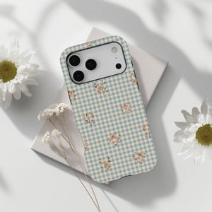 Puede incluir: Una funda de teléfono con un patrón de cuadros vichy azul y crema y pequeños detalles florales. La funda está decorada con un patrón repetitivo de cuadros azul claro y crema, y pequeñas flores color melocotón.