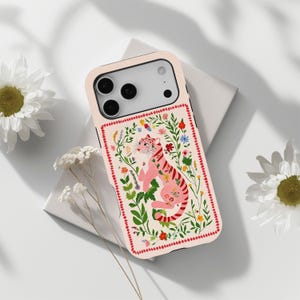 May include: A light pink smartphone case featuring a central illustration of a pink tiger surrounded by colorful wildflowers and green foliage, enclosed within a decorative red dotted border, with cutouts for the camera lenses and flash.