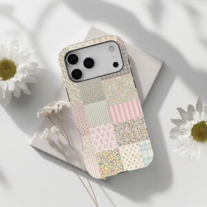 Könnte beinhalten: Handyhülle im Patchwork-Design mit floralen, gepunkteten und gestreiften Mustern in Pastelltönen. Die Hülle hat einen schwarzen Rand und einen Kameraausschnitt. Gänseblümchen und weiße Blumen sind im Hintergrund.