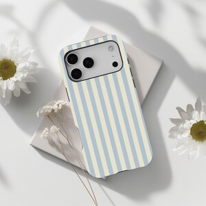 May include: A phone case featuring a light blue and cream vertical stripe design. The case has a camera aperture and is placed on a white surface, accompanied by white flowers.