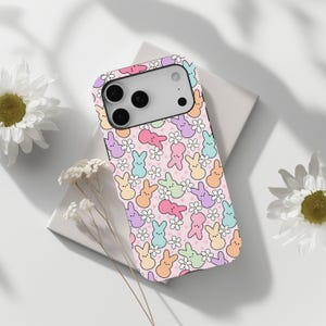 May include: A phone case with a pastel pink background featuring a pattern of colourful cartoon bunnies and white daisy flowers. The case has a black border and a camera cutout.