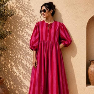 May include: A midi-length dress featuring vertical stripes in alternating shades of dark red and bright pink. The garment has a round neckline with a tie closure, voluminous puff sleeves that reach the elbows, and a gathered waistline.