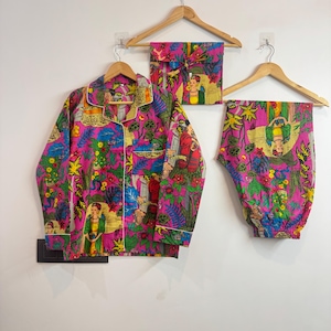 Puede incluir: Un conjunto de pijama rosa con un estampado floral y de figuras colorido. El conjunto incluye una camisa con botones, pantalones y una bolsa a juego, todo colgado en perchas de madera contra una pared blanca. El pijama tiene ribetes blancos.