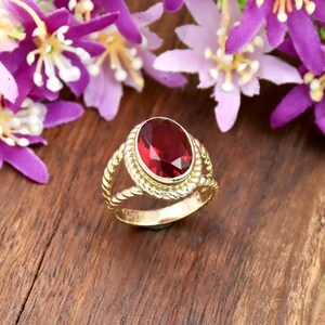 Könnte beinhalten: Ein Goldring mit einem großen, oval geschliffenen, tiefroten Edelstein in einer seilartig texturierten Fassung. Die Ringschiene besteht aus zwei gedrehten Golddrähten, die an der Basis zusammenlaufen und ein geteiltes Schienendesign bilden.