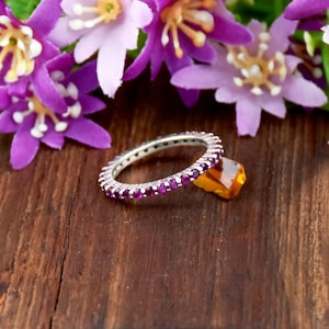 May include: A silver eternity ring featuring a continuous band of small, round, vibrant magenta-pink gemstones. The ring is positioned next to a single, faceted, translucent amber-colored stone, showcasing a clear contrast in color and shape.