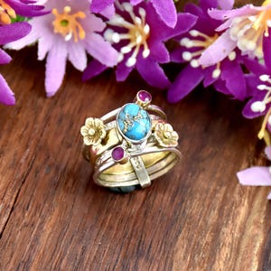 Puede incluir: Un conjunto de anillos apilables con una piedra turquesa central, pequeños detalles de flores doradas y dos pequeñas piedras rojas. Los anillos se muestran sobre una superficie de madera, con flores moradas en el fondo.