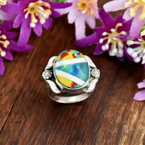 Op de afbeelding: Een zilveren ring met een ovaalvormige inleg met een veelkleurig patroon van gele, blauwe, rode en witte segmenten. De metalen ringband is aan weerszijden van de centrale zetting versierd met kleine bloemmotieven.