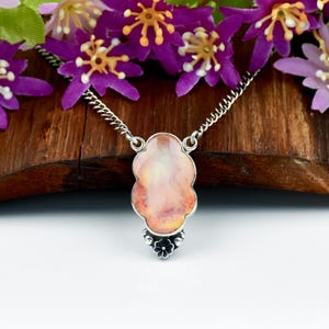 May include: A silver necklace with a pendant featuring a light pink and orange stone. The pendant has a unique, cloud-like shape with a small flower design at the bottom. The necklace is displayed on a wooden surface with purple flowers.