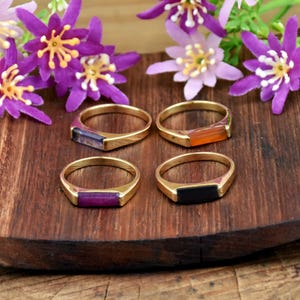 May include: Four gold-toned rings, each featuring a single rectangular gemstone set horizontally. The stones are purple, orange, deep magenta, and black. The rings are arranged in a two-by-two grid on a dark brown wooden surface with purple flowers.
