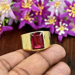 Puede incluir: Un anillo de banda ancha de oro con una gran piedra preciosa rectangular facetada de color rojo intenso engastada en el centro. El metal tiene un acabado liso y pulido, y la piedra muestra un tono carmesí vibrante y translúcido.
