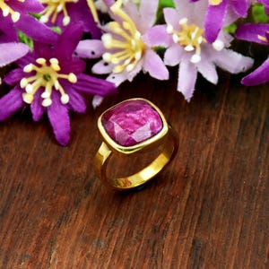 Può includere: Un anello in oro con una pietra preziosa color magenta dal taglio quadrato e sfaccettato, incastonata in una montatura in metallo liscio. L'anello è posizionato su uno sfondo di vivaci fiori viola con centri gialli, creando un contrasto.
