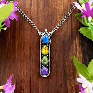 May include: A silver-toned pendant necklace featuring four vertically aligned, teardrop-shaped stones in blue, yellow, green, and purple. The stones are set within a twisted metal frame, suspended from a delicate silver-toned chain link.