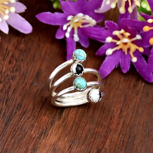 May include: A silver spiral ring featuring four round cabochon stones set in scalloped bezels. The stones include two light blue turquoise-colored gems, one solid black stone, and one solid white stone, arranged along the coiled metal band.