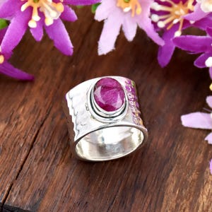 May include: A wide silver ring with a large oval ruby gemstone. The ring has a hammered texture on the band and is set against a wooden surface with purple flowers.