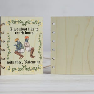 Può includere: Quaderno in legno con illustrazione medievale e la scritta "I wouldst like to touch butts with thee, Valentine!" Il quaderno è rilegato con spago e ha una copertina posteriore vuota.