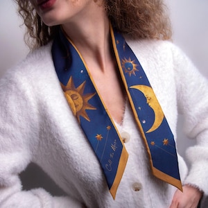 May include: A navy blue scarf with gold trim featuring celestial designs. The scarf displays a sun, moon, stars, and the text "Call the Moon". The scarf is worn over a white cardigan.