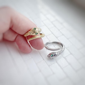 May include: Two snake rings, one gold-toned and one silver-toned, are displayed. The gold ring is held by a finger and features a textured design with green gemstone eyes. The silver ring has a similar snake design with clear stones and green eyes.