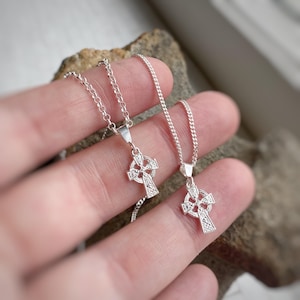 Può includere: Due collane con croci celtiche in argento con intricati disegni a nodi. I ciondoli pendono da delicate catene. Le collane sono esposte su uno sfondo neutro, evidenziando la dettagliata lavorazione artigianale dei gioielli.