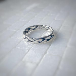 May include: A silver ring with a braided design. The ring is made of a shiny metal and has a textured surface. The ring is on a white surface.