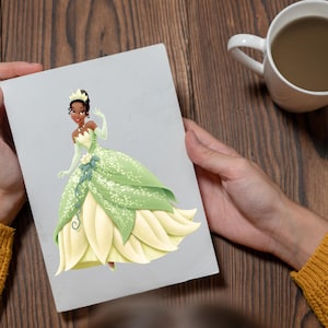 May include: A greeting card featuring a cartoon illustration of a princess in a green and yellow gown. The princess has dark skin, a tiara, and is waving. The card is held in front of a wooden background, with a white mug visible.