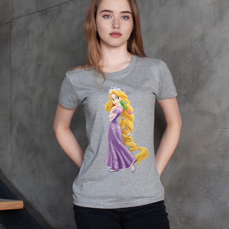 May include: A gray t-shirt featuring a graphic of a cartoon princess with long blonde hair, wearing a purple dress and a crown. The princess is holding a green frog. The shirt has short sleeves and a crew neck.