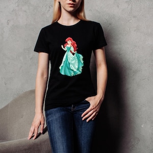 May include: Black t-shirt featuring a graphic of a red-haired princess in a teal gown. The princess is depicted in a flowing dress with a graceful pose. The t-shirt is a classic crew neck style.