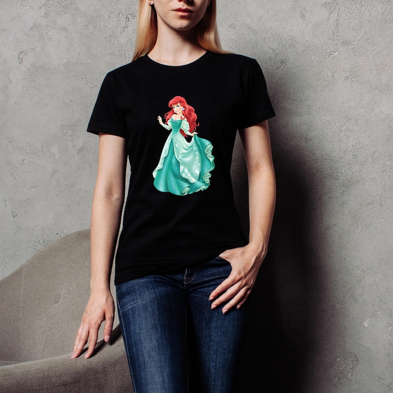 May include: Black t-shirt featuring a graphic of a red-haired princess in a teal gown. The princess is depicted in a flowing dress with a graceful pose. The t-shirt is a classic crew neck style.