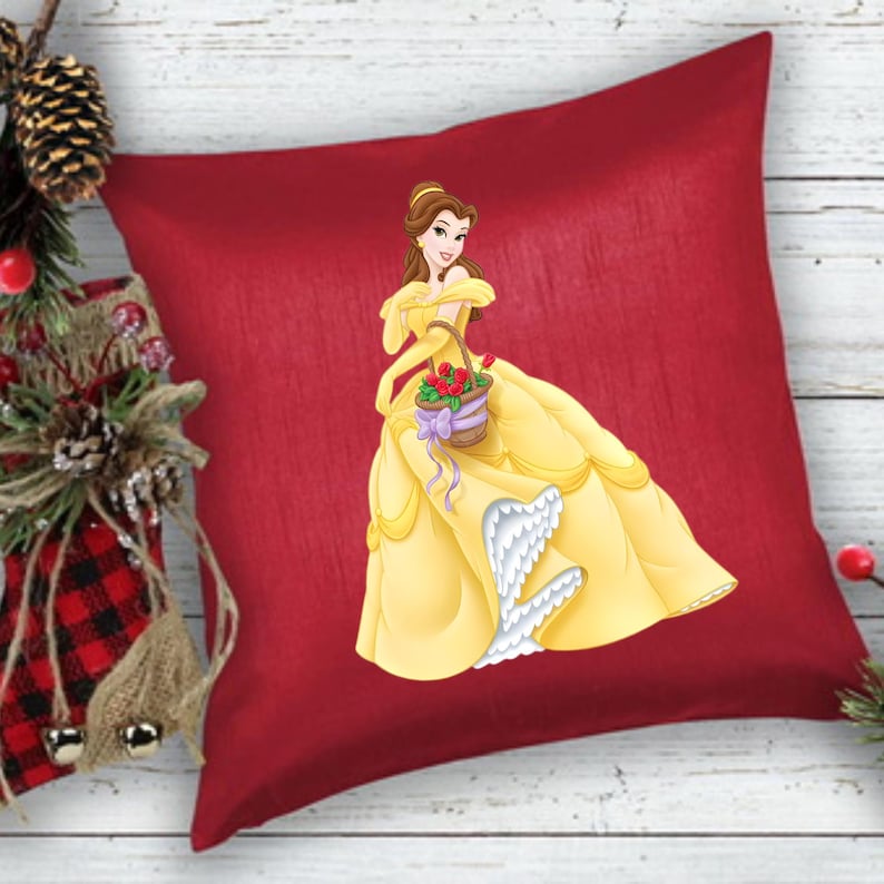 May include: A red decorative pillow featuring a cartoon illustration of a woman in a yellow gown carrying a basket of flowers. The pillow has a solid red background and a textured appearance. The woman is wearing a yellow dress with white accents.