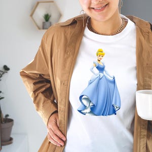 May include: White t-shirt featuring a cartoon illustration of Cinderella in her iconic blue ball gown. The princess is depicted with her signature blonde updo and gloves. The shirt is a classic crew neck style.