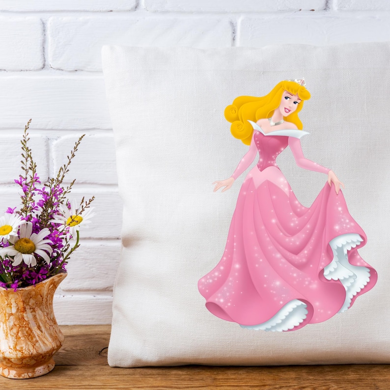May include: A white square pillow featuring a cartoon illustration of a princess in a pink gown with a white trim. The princess has blonde hair and is wearing a crown. The pillow is on a wooden surface with a vase of flowers to the left.