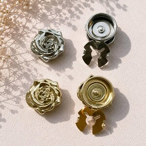 May include: Two metal rose-shaped buttons with snap fasteners. One button is silver-toned and the other is gold-toned. Each button features a detailed, layered rose design on the front and a circular snap mechanism with a pronged base on the back.