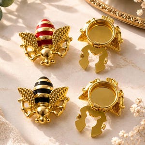 May include: Two bee-shaped metal ornaments with gold-tone bodies and textured wings. One bee features red and gold stripes, while the other has black and gold stripes. Each piece includes a circular, hollow gold-tone backing with a hinged component.
