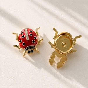 May include: A gold-toned ladybug brooch featuring a vibrant red enamel shell with black spots and a central vertical row of clear crystals. The reverse side shows a circular gold-toned metal backing with a secure clip mechanism for attachment.