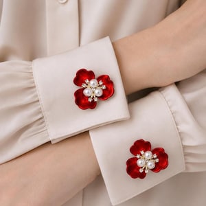 May include: A pair of red flower-shaped cufflinks attached to the cuffs of a beige long-sleeved shirt. Each cufflink features three glossy red petals, a central cluster of three white faux pearls, and small clear crystals set in gold-tone metal.