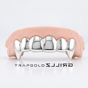 May include: A set of silver-colored vampire fang grillz fitted over a light pink gum model. The metallic teeth feature pointed canine fangs on each end. The text TRAPGOLD GRILLZ is printed in black below the dental appliance on a white background.