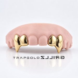 May include: A set of gold teeth grillz featuring pointed canine caps on a light pink dental model. The gold caps are polished and reflective. The text TRAPGOLD GRILLZ is printed in black below the dental model on a white surface.