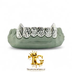 May include: A set of silver-colored grillz with a geometric pattern, displayed on a gray dental mold. The grillz feature a detailed, textured design. The logo "TRAPGOLDGRILLZ - CUSTOM JEWELRY FINE" is at the bottom.