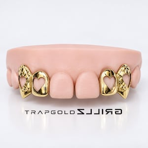 May include: Gold grillz set featuring four teeth covers with heart-shaped cutouts and diamond-cut textured detailing on the outer teeth. The gold teeth are displayed on a light pink dental model. The text TRAPGOLDZ is printed below.