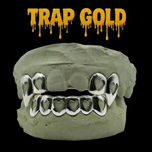 May include: Silver-colored grillz with heart-shaped cutouts, displayed on a light green dental mold. The text "TRAP GOLD" is written in gold dripping font above the grillz against a black background.