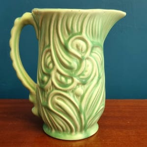 May include: A light green ceramic pitcher with a decorative floral pattern. The pitcher has a curved handle and a spout for pouring. The design features stylized leaves and swirls, with a glossy finish. The pitcher is a decorative item.