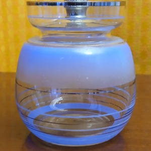 May include: A vintage glass jar with a silver metal lid and accents. The jar has a light blue frosted band and is decorated with blue and black spiral lines. The jar is likely used for storing small items or as a decorative piece.