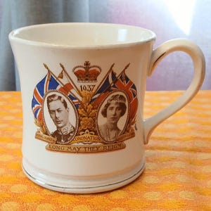 May include: Cream-colored ceramic mug featuring portraits of a man and woman, flags, and a crown. The mug has a gold rim and handle, with the text "1937 Coronation" and "Long May They Reign".