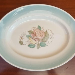 May include: A white oval serving platter with a light blue rim and a floral design featuring a pink rose with green leaves.